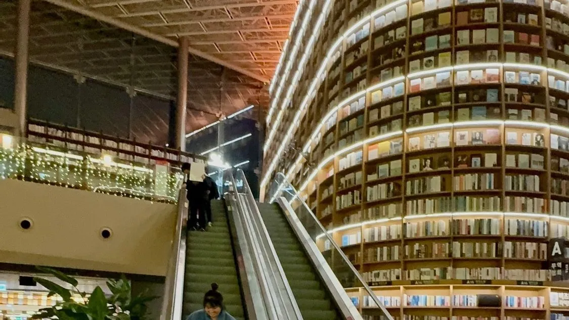 Escalators rising past a curved, illuminated bookshelf tower that climbs to the mall ceiling